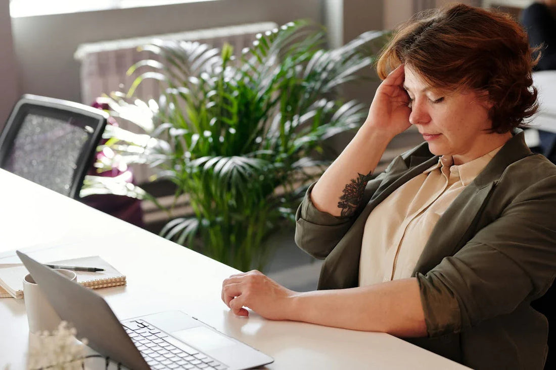 Vrouw met hand aan hoofd werkt gestrest op kantoor aan laptop, planten op achtergrond