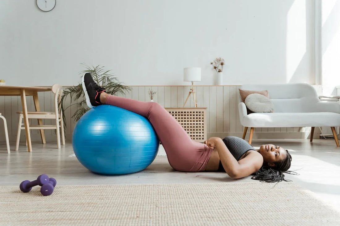 Vrouw doet fitnessoefening voor spiermassa met blauwe fitnessbal in woonkamer, trainingsmat en halters