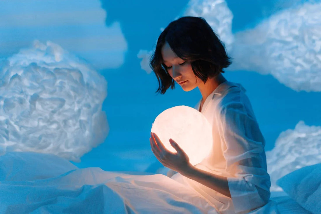 Vrouw in wit nachtkleed houdt gloeilamp maan vast tussen nepwolken in blauwe kamer
